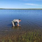 Mehdi stands in a lake with his hand under water wearing waders